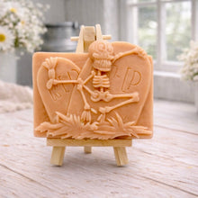 Load image into Gallery viewer, A handmade vegan soap depicting a skeleton resting between two R.I.P. gravestones, displayed on a small wooden easel.