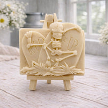 Load image into Gallery viewer, A handmade vegan soap depicting a skeleton resting between two R.I.P. gravestones, displayed on a small wooden easel.
