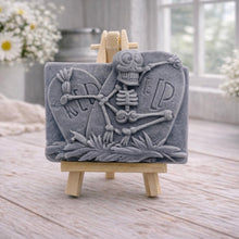 Load image into Gallery viewer, A handmade vegan soap depicting a skeleton resting between two R.I.P. gravestones, displayed on a small wooden easel.