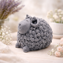 Load image into Gallery viewer, A set of three handmade vegan soaps in different sheep styles, including a dark grey smiling sheep and two lambs with detailed woolly textures, displayed on a rustic wooden surface with lavender and flowers.