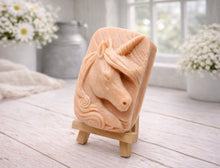 Load image into Gallery viewer, handmade vegan soap featuring a detailed unicorn head relief with a spiralled horn and flowing curly mane, displayed on a small wooden easel in a bright farmhouse decor setting.