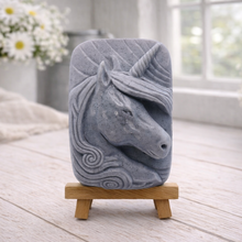 Load image into Gallery viewer, handmade vegan soap featuring a detailed unicorn head relief with a spiralled horn and flowing curly mane, displayed on a small wooden easel in a bright farmhouse decor setting.