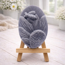 Load image into Gallery viewer, A handmade vegan soap in a natural colour, featuring an oval relief of a honeybee landing on a large sunflower with detailed petals and a textured centre, displayed on a small wooden easel.
