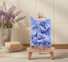 Load image into Gallery viewer, A rectangular handmade vegan soap, featuring a high-relief carving of a seated fairy with large, detailed wings holding a single blossom, displayed on a miniature wooden easel.