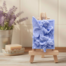 Load image into Gallery viewer, A rectangular handmade vegan soap, featuring a high-relief carving of a seated fairy with large, detailed wings holding a single blossom, displayed on a miniature wooden easel.