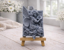 Load image into Gallery viewer, A rectangular handmade vegan soap, featuring a high-relief carving of a seated fairy with large, detailed wings holding a single blossom, displayed on a miniature wooden easel.