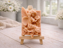Load image into Gallery viewer, A rectangular handmade vegan soap, featuring a high-relief carving of a seated fairy with large, detailed wings holding a single blossom, displayed on a miniature wooden easel.