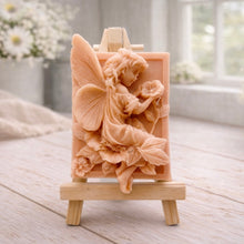 Load image into Gallery viewer, A rectangular handmade vegan soap, featuring a high-relief carving of a seated fairy with large, detailed wings holding a single blossom, displayed on a miniature wooden easel.