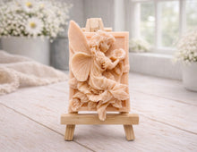 Load image into Gallery viewer, A rectangular handmade vegan soap, featuring a high-relief carving of a seated fairy with large, detailed wings holding a single blossom, displayed on a miniature wooden easel.