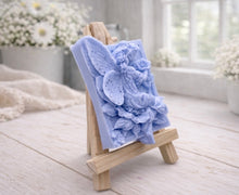 Load image into Gallery viewer, A rectangular handmade vegan soap, featuring a high-relief carving of a seated fairy with large, detailed wings holding a single blossom, displayed on a miniature wooden easel.
