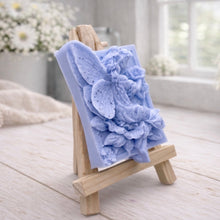 Load image into Gallery viewer, A rectangular handmade vegan soap, featuring a high-relief carving of a seated fairy with large, detailed wings holding a single blossom, displayed on a miniature wooden easel.