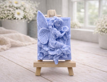 Load image into Gallery viewer, A rectangular handmade vegan soap, featuring a high-relief carving of a seated fairy with large, detailed wings holding a single blossom, displayed on a miniature wooden easel.