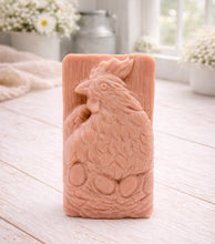 Load image into Gallery viewer, A rectangular handmade vegan soap in a natural colour, featuring a highly detailed relief carving of a broody hen chicken sitting on a nest of eggs, displayed upright on a light wooden surface.