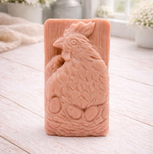 Load image into Gallery viewer, A rectangular handmade vegan soap in a natural colour, featuring a highly detailed relief carving of a broody hen chicken sitting on a nest of eggs, displayed upright on a light wooden surface.