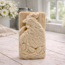 Load image into Gallery viewer, A rectangular handmade vegan soap in a natural colour, featuring a highly detailed relief carving of a broody hen chicken sitting on a nest of eggs, displayed upright on a light wooden surface.