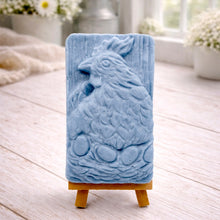 Load image into Gallery viewer, A rectangular handmade vegan soap in a natural colour, featuring a highly detailed relief carving of a broody hen chicken sitting on a nest of eggs, displayed upright on a light wooden surface.