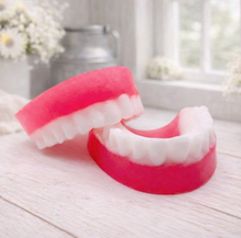 Load image into Gallery viewer, A 3D handmade vegan soap sculpted to look like a realistic set of dentures, featuring vibrant pink gums and a full row of white teeth, resting on a light wooden surface in a bright farmhouse setting.