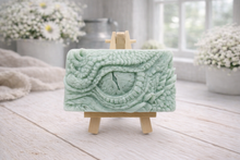 Load image into Gallery viewer, A rectangular handmade vegan soap in a slate-grey tone, featuring a highly detailed, close-up relief of a mythical dragon or dinosaur eye surrounded by thick, pebbled scales and reptilian skin textures, displayed on a miniature wooden easel.