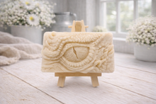 Load image into Gallery viewer, A rectangular handmade vegan soap in a slate-grey tone, featuring a highly detailed, close-up relief of a mythical dragon or dinosaur eye surrounded by thick, pebbled scales and reptilian skin textures, displayed on a miniature wooden easel.