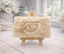 Load image into Gallery viewer, A rectangular handmade vegan soap in a slate-grey tone, featuring a highly detailed, close-up relief of a mythical dragon or dinosaur eye surrounded by thick, pebbled scales and reptilian skin textures, displayed on a miniature wooden easel.
