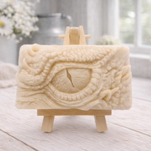 Load image into Gallery viewer, A rectangular handmade vegan soap in a slate-grey tone, featuring a highly detailed, close-up relief of a mythical dragon or dinosaur eye surrounded by thick, pebbled scales and reptilian skin textures, displayed on a miniature wooden easel.
