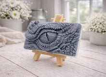 Load image into Gallery viewer, A rectangular handmade vegan soap in a slate-grey tone, featuring a highly detailed, close-up relief of a mythical dragon or dinosaur eye surrounded by thick, pebbled scales and reptilian skin textures, displayed on a miniature wooden easel.