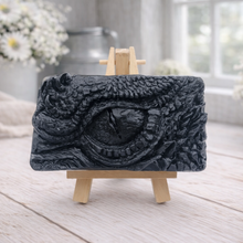 Load image into Gallery viewer, A rectangular handmade vegan soap in a slate-grey tone, featuring a highly detailed, close-up relief of a mythical dragon or dinosaur eye surrounded by thick, pebbled scales and reptilian skin textures, displayed on a miniature wooden easel.