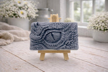 Load image into Gallery viewer, A rectangular handmade vegan soap in a slate-grey tone, featuring a highly detailed, close-up relief of a mythical dragon or dinosaur eye surrounded by thick, pebbled scales and reptilian skin textures, displayed on a miniature wooden easel.