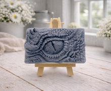 Load image into Gallery viewer, A rectangular handmade vegan soap in a slate-grey tone, featuring a highly detailed, close-up relief of a mythical dragon or dinosaur eye surrounded by thick, pebbled scales and reptilian skin textures, displayed on a miniature wooden easel.