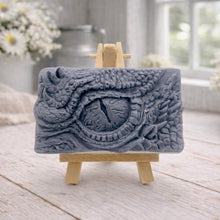 Load image into Gallery viewer, A rectangular handmade vegan soap in a slate-grey tone, featuring a highly detailed, close-up relief of a mythical dragon or dinosaur eye surrounded by thick, pebbled scales and reptilian skin textures, displayed on a miniature wooden easel.