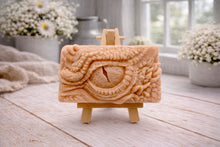 Load image into Gallery viewer, A rectangular handmade vegan soap in a slate-grey tone, featuring a highly detailed, close-up relief of a mythical dragon or dinosaur eye surrounded by thick, pebbled scales and reptilian skin textures, displayed on a miniature wooden easel.