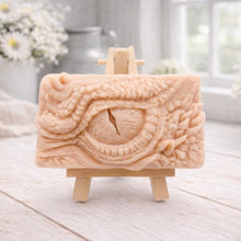 Load image into Gallery viewer, A rectangular handmade vegan soap in a slate-grey tone, featuring a highly detailed, close-up relief of a mythical dragon or dinosaur eye surrounded by thick, pebbled scales and reptilian skin textures, displayed on a miniature wooden easel.