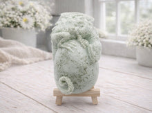 Load image into Gallery viewer, A 3D handmade vegan soap in a charcoal-grey tone, sculpted as a large prehistoric egg with a small dinosaur or dragon clinging to the top, featuring detailed scales and a curled tail, displayed on a miniature wooden easel.