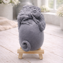 Load image into Gallery viewer, A 3D handmade vegan soap in a charcoal-grey tone, sculpted as a large prehistoric egg with a small dinosaur or dragon clinging to the top, featuring detailed scales and a curled tail, displayed on a miniature wooden easel.