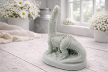 Load image into Gallery viewer, A handmade vegan soap in the shape of a long-necked brachiosaurus dinosaur, featuring a soft mint green colour with natural speckles, set against a bright window and white flowers