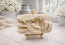 Load image into Gallery viewer, A rectangular handmade vegan soap featuring a high-relief carving of a Tyrannosaurus Rex with its jaws wide open in a roar and sharp claws framing the face, displayed on a miniature wooden easel.