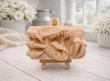 Load image into Gallery viewer, A rectangular handmade vegan soap featuring a high-relief carving of a Tyrannosaurus Rex with its jaws wide open in a roar and sharp claws framing the face, displayed on a miniature wooden easel.