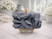 Load image into Gallery viewer, A rectangular handmade vegan soap in a charcoal-grey tone, featuring a high-relief carving of a Tyrannosaurus Rex with its jaws wide open in a roar and sharp claws framing the face, displayed on a miniature wooden easel.