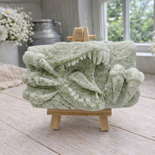 Load image into Gallery viewer, A rectangular handmade vegan soap featuring a high-relief carving of a Tyrannosaurus Rex with its jaws wide open in a roar and sharp claws framing the face, displayed on a miniature wooden easel.