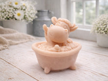 Load image into Gallery viewer, A handmade vegan soap is sculpted in the shape of a "dirty cow" character happily sitting in a bubble-filled roll-top bathtub with one hoof raised, displayed on a light wooden surface.