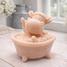 Load image into Gallery viewer, A handmade vegan soap is sculpted in the shape of a "dirty cow" character happily sitting in a bubble-filled roll-top bathtub with one hoof raised, displayed on a light wooden surface.