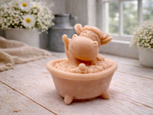 Load image into Gallery viewer, A handmade vegan soap is sculpted in the shape of a "dirty cow" character happily sitting in a bubble-filled roll-top bathtub with one hoof raised, displayed on a light wooden surface.