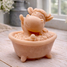 Load image into Gallery viewer, A handmade vegan soap is sculpted in the shape of a "dirty cow" character happily sitting in a bubble-filled roll-top bathtub with one hoof raised, displayed on a light wooden surface.