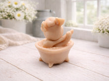 Load image into Gallery viewer, A handmade vegan soap is sculpted in the shape of a "dirty cow" character happily sitting in a bubble-filled roll-top bathtub with one hoof raised, displayed on a light wooden surface.