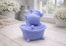 Load image into Gallery viewer, A handmade vegan soap is sculpted in the shape of a "dirty cow" character happily sitting in a bubble-filled roll-top bathtub with one hoof raised, displayed on a light wooden surface.