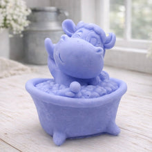 Load image into Gallery viewer, A handmade vegan soap is sculpted in the shape of a "dirty cow" character happily sitting in a bubble-filled roll-top bathtub with one hoof raised, displayed on a light wooden surface.