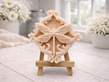 Load image into Gallery viewer, A beautiful handmade vegan soap in a natural tone, featuring a highly detailed relief of a dragonfly with delicate, outspread wings and a textured body, displayed on a miniature wooden easel against a soft farmhouse background.