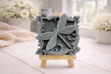 Load image into Gallery viewer, A beautiful handmade vegan soap in a natural tone, featuring a highly detailed relief of a dragonfly with delicate, outspread wings and a textured body, displayed on a miniature wooden easel against a soft farmhouse background.