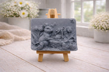 Load image into Gallery viewer, A rectangular handmade vegan soap in a sophisticated slate-grey tone, featuring a highly detailed relief scene of an Easter bunny standing next to a basket of eggs, surrounded by blooming tulips and small birds, displayed on a wooden easel.