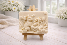 Load image into Gallery viewer, A rectangular handmade vegan soap in a sophisticated slate-grey tone, featuring a highly detailed relief scene of an Easter bunny standing next to a basket of eggs, surrounded by blooming tulips and small birds, displayed on a wooden easel.